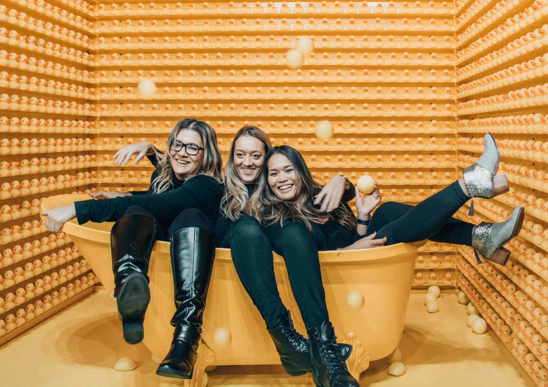 Three women posing in a yellow bathtub with rubber duck walls.